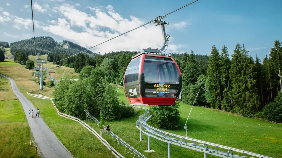 Rote Seilbahngondel über grüner Hügellandschaft mit Wald im Hintergrund.