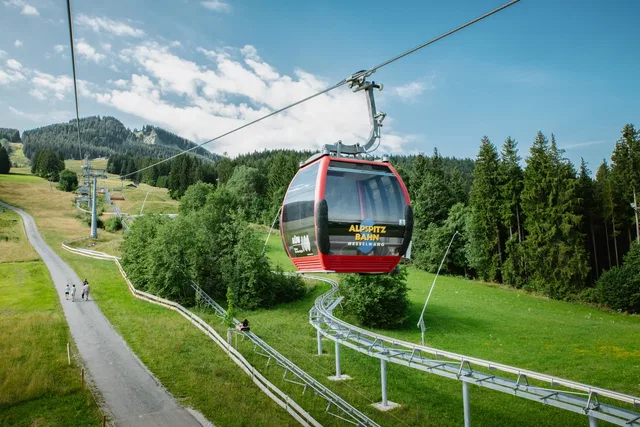 Rote Seilbahngondel über grüner Hügellandschaft mit Wald im Hintergrund.