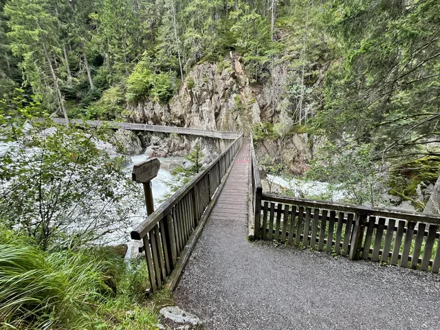 Holzbrücke über Fluss, umgeben von dichtem Wald und Felswänden.