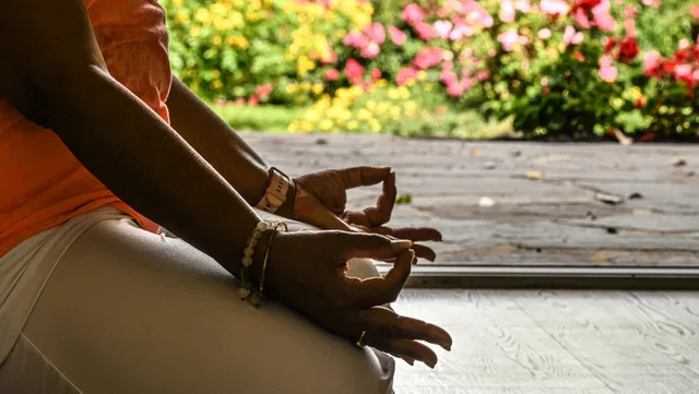 Person sitzt im Lotus-Sitz, Hände in Mudra, farbenfrohe Blumen im Hintergrund.