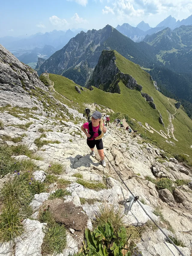 Frau wandert steil auf felsigem Grat in den Alpen, weitere Gruppenwanderer im Hintergrund.