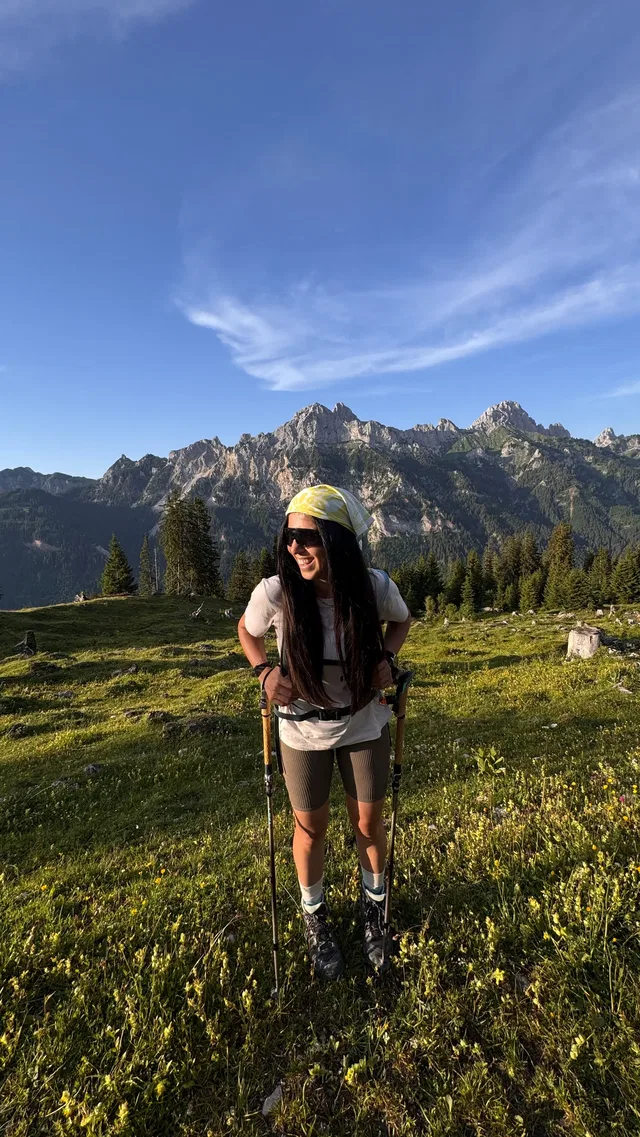 Frau mit Wanderstöcken in einer grünen Bergwiese vor felsigen Gipfeln.