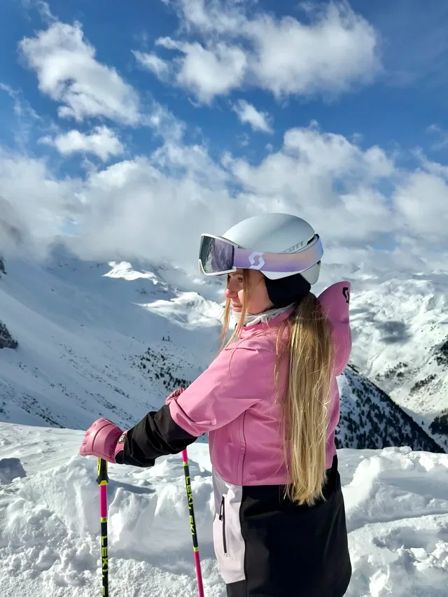 Skifahrerin in pinker Jacke mit Helm und Skibrille vor verschneiter Berglandschaft.