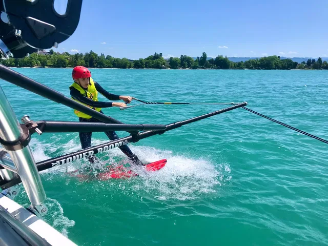 Wakeboarder wird von einem Boot mit Seil über das Wasser gezogen, roter Helm, gelbe Rettungsweste.