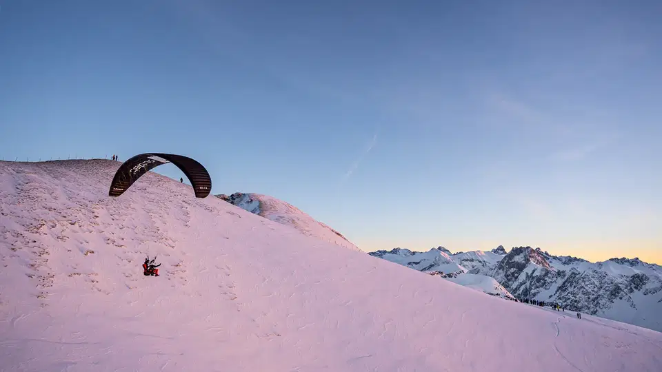 Schwarzer Gleitschirm beim Start an einem verschneiten Hang im Abendlicht.